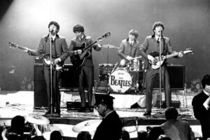 Foto histórica em preto e branco dos Beatles se apresentando ao vivo no palco, representando a Beatlemania, o início do rock e a invasão britânica nos anos 60.